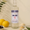 In der Mitte steht eine Flasche Ouzo und vor einer Wand. Neben dran liegen dekoartikel wie eine Zintrone und Oliven.