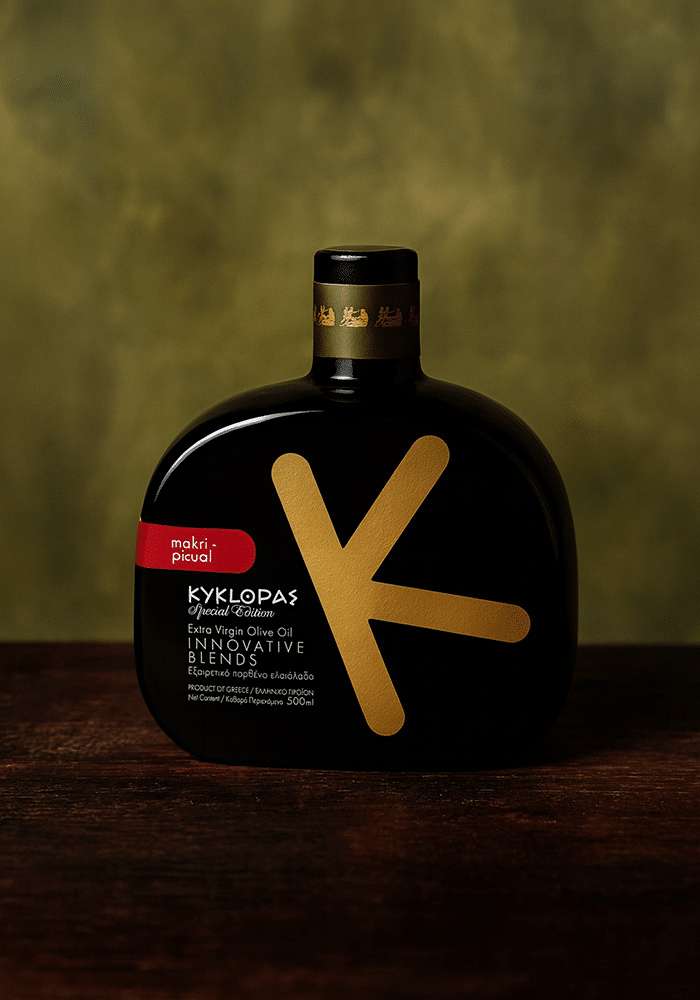 Schwarze, flache Flasche mit goldfarbenem „K“-Design und Aufschrift „KYKLOPAS Special Edition Extra Virgin Olive Oil Innovative Blends, Makri-Picual“ auf dunklem Holz vor verschwommenem, olivgrünem Hintergrund.